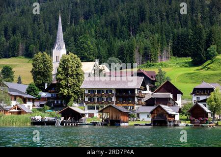 Schöne traditionelle Holzhäuser über dem Wasser an einem spektakulären Weissensee in Kärnten in Österreich, mit Hotels und Kirche im Hintergrund Stockfoto