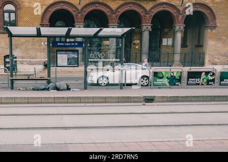 Ein Obdachloser schläft an der Straßenbahnhaltestelle. In Ungarn, insbesondere in Budapest, herrscht anhaltende Besorgnis über Obdachlosigkeit. Aufgrund der hohen Inflation ist die Zahl der Menschen, die auf den Straßen leben, in der Stadt gestiegen. Faktoren wie Armut, Arbeitslosigkeit, psychische Gesundheit und Mangel an bezahlbarem Wohnraum tragen zu dem Problem bei. Die ungarische Regierung und die lokalen Behörden haben verschiedene Maßnahmen zur Bekämpfung der Obdachlosigkeit ergriffen. Dazu gehören die Bereitstellung von Notunterkünften, soziale Dienste und Programme zur Wiedereingliederung in die Gesellschaft. Nichtregierungsorganisationen oder Stockfoto