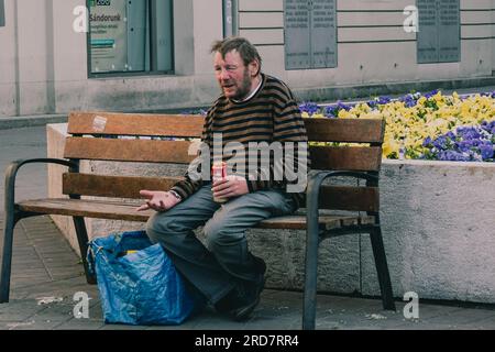 Ein Obdachloser sitzt auf einer Bank und trinkt ein Bier. In Ungarn, insbesondere in Budapest, herrscht anhaltende Besorgnis über Obdachlosigkeit. Aufgrund der hohen Inflation ist die Zahl der Menschen, die auf den Straßen leben, in der Stadt gestiegen. Faktoren wie Armut, Arbeitslosigkeit, psychische Gesundheit und Mangel an bezahlbarem Wohnraum tragen zu dem Problem bei. Die ungarische Regierung und die lokalen Behörden haben verschiedene Maßnahmen zur Bekämpfung der Obdachlosigkeit ergriffen. Dazu gehören die Bereitstellung von Notunterkünften, soziale Dienste und Programme zur Wiedereingliederung in die Gesellschaft. Nein Stockfoto