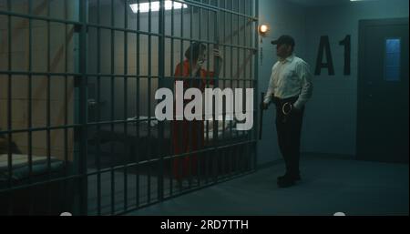 Ein Gefängniswärter mit Polizeistab steht in der Nähe der Gefängniszelle, spricht mit einer Gefangenen in orangefarbener Uniform durch Metallgitter. Frauen sitzen im Gefängnis. Haftanstalt oder Strafvollzugsanstalt. Stockfoto