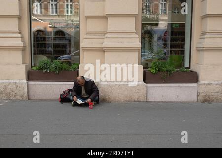 3. Mai 2023, Budapest, Ungarn: Ein Obdachloser, der vor einem Geschäft ein Buch lesen sah. In Ungarn, insbesondere in Budapest, herrscht anhaltende Besorgnis über Obdachlosigkeit. Aufgrund der hohen Inflation ist die Zahl der Menschen, die auf den Straßen leben, in der Stadt gestiegen. Faktoren wie Armut, Arbeitslosigkeit, psychische Gesundheit und Mangel an bezahlbarem Wohnraum tragen zu dem Problem bei. Die ungarische Regierung und die lokalen Behörden haben verschiedene Maßnahmen zur Bekämpfung der Obdachlosigkeit ergriffen. Dazu gehören die Bereitstellung von Notunterkünften, soziale Dienste und Programme, die auf Reinteg abzielen Stockfoto