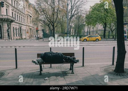 10. April 2023, Budapest, Ungarn: Ah Obdachloser, der auf einer Bank in der Nähe der Straße schläft. In Ungarn, insbesondere in Budapest, herrscht anhaltende Besorgnis über Obdachlosigkeit. Aufgrund der hohen Inflation ist die Zahl der Menschen, die auf den Straßen leben, in der Stadt gestiegen. Faktoren wie Armut, Arbeitslosigkeit, psychische Gesundheit und Mangel an bezahlbarem Wohnraum tragen zu dem Problem bei. Die ungarische Regierung und die lokalen Behörden haben verschiedene Maßnahmen zur Bekämpfung der Obdachlosigkeit ergriffen. Dazu gehören die Bereitstellung von Notunterkünften, Sozialdienstleistungen und Programme für RE Stockfoto