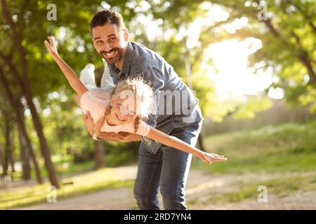 Aufgeregtes kleines Mädchen, das mit ihrem glücklichen Vater mittleren Alters im Park spielt, Kind streckt sich die Hände, gibt vor zu fliegen Stockfoto