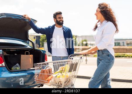 Ein glücklicher araber, der den Kofferraum öffnet, um Produkte zu packen, eine Frau, die mit einem Einkaufswagen voller frischer Nahrung steht Stockfoto