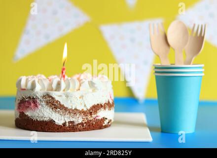 Festlicher Kuchen mit einer Geburtstagskerze. Kuchen und Pappbecher mit Löffeln und Gabeln. Kuchen auf festlichem Hintergrund. Herzlichen Glückwunsch Stockfoto