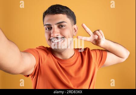 Ein sorgenloser junger brasilianischer Mann, der Selfie macht, friedliche Geste zeigt und lächelt, verspielte Stimmung hat, orangefarbener Hintergrund Stockfoto