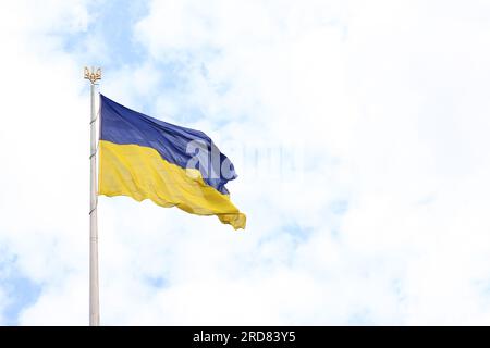 Die Flagge und das Wappen der Ukraine auf einem Flaggenmast vor einem wolkigen Himmel. Die ukrainische Flagge entwickelt sich im Wind Stockfoto