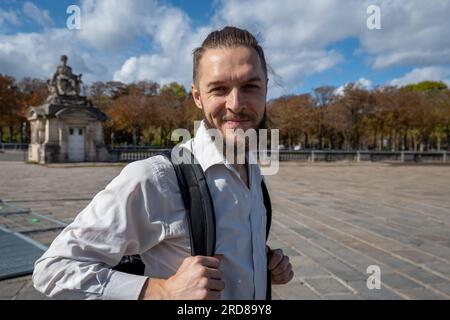 Weißer junger bärtiger männlicher Tourist in einem weißen Hemd in Paris mit einem Lächeln im Rucksack in der Kamera in der Nähe des Triumphbogens Stockfoto