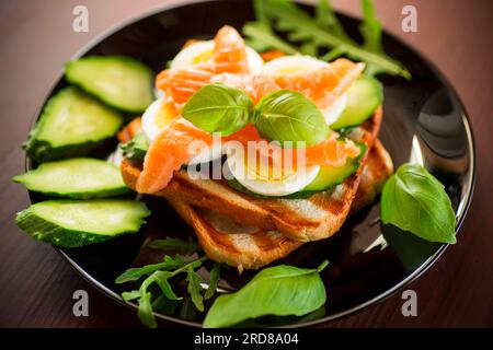 Gekochtes Sandwich mit leicht gesalzenem Rotfisch, Gurken und gekochten Eiern mit Kräutern, auf einem Teller. Stockfoto