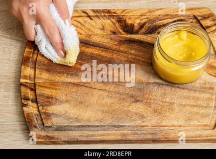 Frauenhände tragen hausgemachtes Bienenwachs-Holzpolitur auf, um das Naturholz-Schneidebrett wiederherzustellen. Bienenwachs, Olivenöl und ätherisches Öl, weiches Tuch. Stockfoto