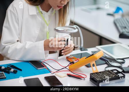 Zugeschnittenes Bild des Serviceleiters mit Multimeter bei der Untersuchung des Smartphones eines Kunden Stockfoto