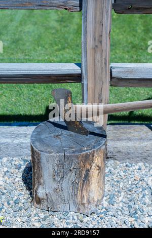 Axt, die einen Holzstumpf schneidet oder auf einem Holzstumpf ruht. Die Axt hat eine rostige Klinge und einen Holzgriff. Ein Zaun ist in der Nähe im Hof. Stockfoto