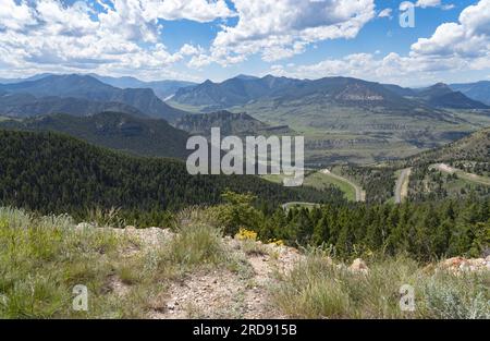 Von diesem Aussichtspunkt abseits des Highways sehen Sie die Straße, die sich zum Sunlight Basin im Norden von Wyoming schlängelt. Wolken verbinden sich mit blauem Himmel. Berggipfel gehen für immer. Stockfoto