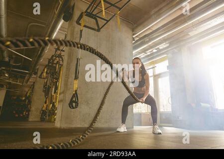 Eine sportliche Frau trainiert Trizeps mit zwei Kabeln an ihren Händen auf dem Hintergrund des Fitnesscenters Stockfoto