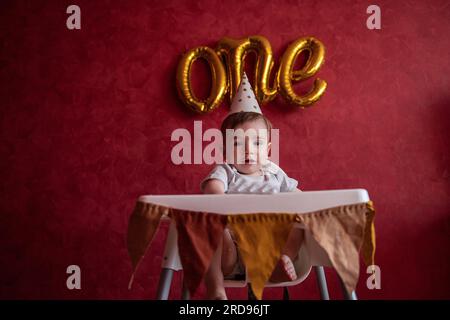 Portrait eines einjährigen Jungen mit festlichem Hut sitzt auf einem Kinderstuhl mit Stofffahnen. Kind auf isoliertem roten Hintergrund mit goldenen Luftballons aus Folie Stockfoto