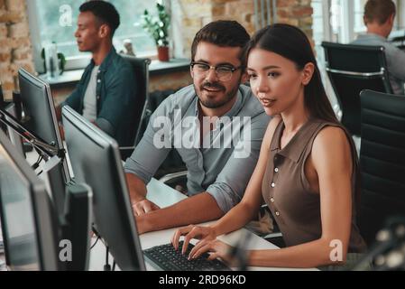 Ich erkläre es Ihnen. Zwei positive junge Menschen schauen auf einen Computermonitor und diskutieren über Arbeitsaufgaben, während ihre Kollegen im Hintergrund arbeiten Stockfoto