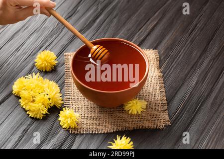 Honig in einer Tonschüssel, Dipper und gelbe Blumen auf Textil- und dunklem Holzhintergrund. Ein Honigstock liegt in einer Keramikschüssel mit Honig Stockfoto