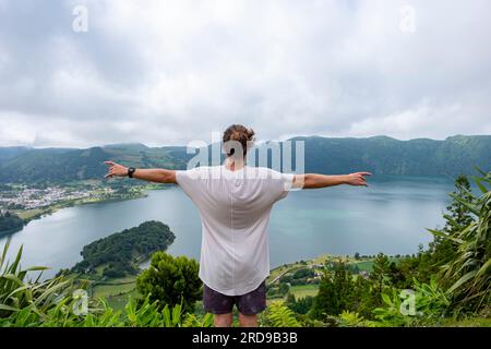 Junger Mann in weißem T-Shirt mit ausgestreckten Armen und Blick auf den See von Seven Cities Lake „Lagoa das Sete Cidades“. Die Insel Sao Miguel auf den Azoren. Stockfoto