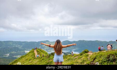 Junge Frau mit lockigem Haar und ausgestreckten Armen, die auf dem Gipfel des Berges steht und auf den See Sete Cidades blickt, vom Aussichtspunkt Grota do Inf Stockfoto