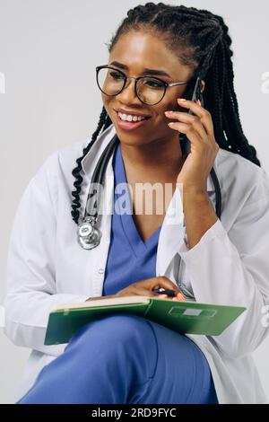 Porträt einer afroamerikanischen Ärztin oder Krankenschwester, die einen Laborkittel trägt und ein Smartphone vor weißem Hintergrund hält Stockfoto