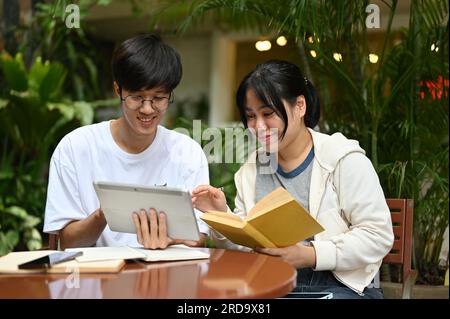 Zwei glückliche junge asiatische Studenten sitzen in einem Café im Außenbereich und arbeiten gemeinsam an einem Schulprojekt oder Hausaufgaben. Stockfoto