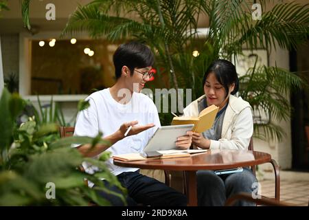 Zwei glückliche junge asiatische Studenten sitzen in einem Café in einem Außenbereich, diskutieren über ihre Arbeit und arbeiten gemeinsam an einem Endprojekt. Stockfoto