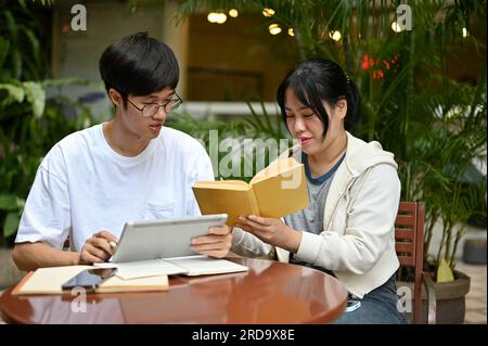 Zwei kluge und fokussierte junge asiatische Studenten sitzen in einem Café in einem Außenbereich, lesen ein Buch und bereiten sich gemeinsam auf die Prüfung vor. Stockfoto