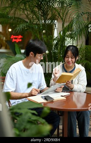 Zwei glückliche junge asiatische Studenten sitzen in einem Café in einem Außenbereich, lesen ein Buch und bereiten sich gemeinsam auf die Prüfung vor. Freundschaft und E Stockfoto