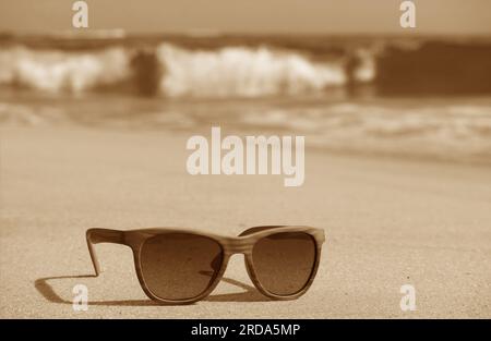 Sepia-Bild einer Sonnenbrille am Sandstrand mit Wellen im Hintergrund Stockfoto