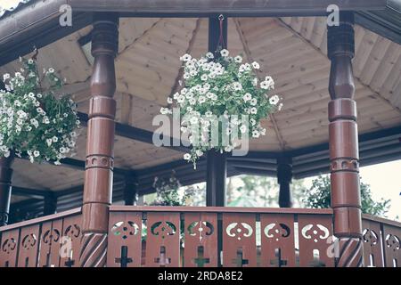 Wunderschöne hölzerne Gartenlaube, dekoriert mit Blumen an einem sonnigen Sommertag. Nahaufnahme eines großen hölzernen Pavillons, mit wunderschönen hängenden Pflanzen, in einem öffentlichen Park. Hochwertiges Foto Stockfoto