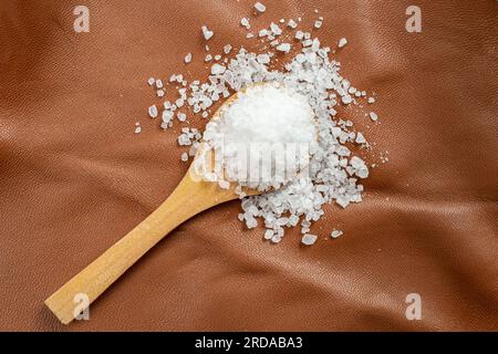 Weißes Steinsalz füllt einen hölzernen Teelöffel auf braunem Leder, Hautstruktur, abstrakter Hintergrund mit weichem Fokus Stockfoto