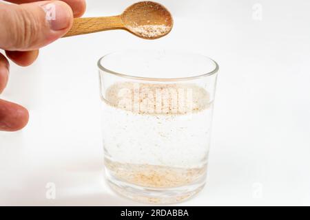 Mischen Sie mit der Hand einen Teelöffel voller Psylliumschalen mit Wasser in einem transparenten Glas auf weißem Hintergrund, Nahaufnahme mit weichem Fokus Stockfoto