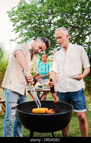 Vertikales Porträt eines Mannes mittleren Alters, der Fleisch auf einer guten Grillparty zuhause kocht. Zwei gut aussehende Freunde oder die Familie grillen auf einem barbecue-Mittagessen und haben Spaß zusammen. Hochwertiges Foto Stockfoto