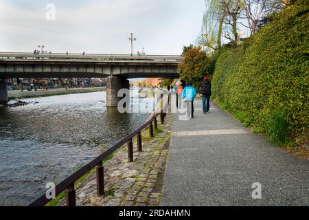 Touristen laufen am Flussufer entlang, Brücke über den Kamo River und Kirschblüte im Hintergrund. Präfektur Kyoto. Japan. Stockfoto
