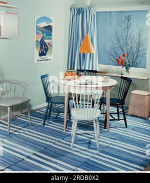Küche in der Vergangenheit. Eine Küche mit einem runden Küchentisch mit vier Küchenstühlen. Auf dem Boden befinden sich handgewebte Teppiche aus blau-weißem Stoff. Eine gelbe Küchenlampe hängt über dem Tisch. An der Wand hängt ein klassisches Regal aus Schnur. Schweden 1958. Conard Ref BV98-6 Stockfoto