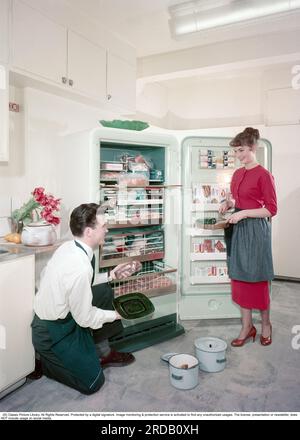 Küche aus dem 1950er. Jahrhundert. Das junge schwedische Paar in der Stilküche. Die Tür zum neuen Gefrierschrank, in dem die Regale mit Tiefkühlwaren gefüllt sind, ist offen. Tiefgefrorene Lebensmittel wären der Beginn einer Revolution der Haushaltsgewohnheiten und der Lebensmittellagerung. Viele waren skeptisch, und insbesondere Hersteller von Konserven betrachteten Tiefkühlkost als vorübergehenden Trend. Schweden 1959. ref. BV111-3 Stockfoto