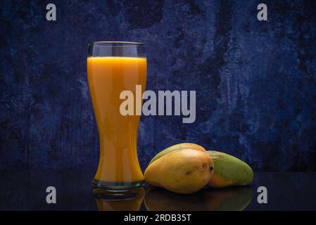 Mangosaftglas und eine Gruppe Mangos auf einem schwarzen Glastisch. Blauer Hintergrund. Stockfoto