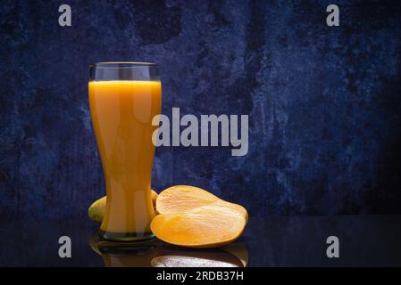 Mangosaftglas und eine Gruppe Mangos auf einem schwarzen Glastisch. Blauer Hintergrund. Stockfoto