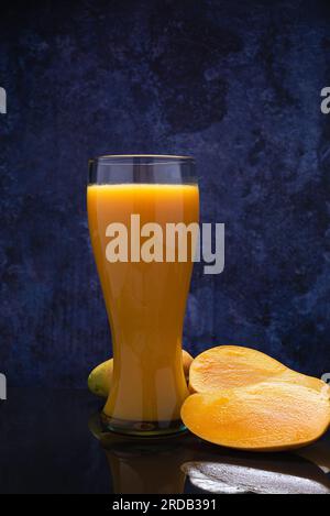 Mangosaftglas und eine Gruppe Mangos auf einem schwarzen Glastisch. Blauer Hintergrund. Stockfoto