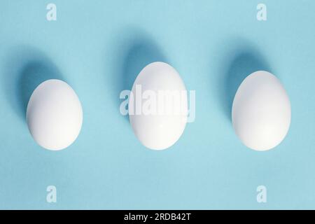 Drei einzigartige weiße Hühnereier auf blauem Hintergrund, Draufsicht. Frische, rohe, ovale Tiereier, Schatten. Natürliche ganze Eierschalen unterschiedlicher Größe in Kuppeln Stockfoto