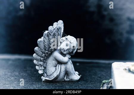 Alte, schmutzige Statue des Traubenengels auf Grabstein auf dem alten Friedhof. Kleine Engelsfigur mit Flügeln auf einem dunklen Traurigkeitsgräber. Baby-Engel-Figur Stockfoto