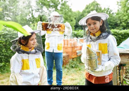 Lächelnde Mädchen mit Raucher stehen neben einem männlichen Imker und halten Wabenrahmen im Garten Stockfoto