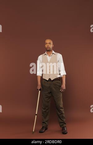Echte Menschen, kühner afroamerikanischer Mann mit Myasthenia-gravis-Syndrom, der mit einem Gehstock auf braunem Hintergrund steht, steht und in die Kamera schaut, Stockfoto