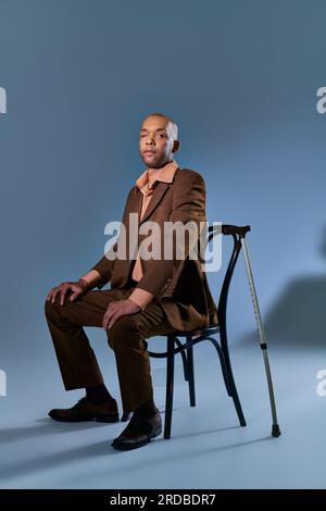 Vielfalt und Inklusion, afroamerikanischer Mann mit Myasthenia-gravis-Syndrom, sitzt auf dem Stuhl und sieht auf blauem Hintergrund vor die Kamera, Gehstock, Stockfoto