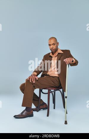 Ethnisch, voller Länge, ein mutiger afroamerikanischer Mann mit Myasthenia gravis, der auf einem Stuhl auf grauem Hintergrund sitzt, dunkelhäutige Person im Anzug, die sich auf wa lehnt Stockfoto