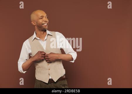 Echte Menschen, fröhlicher und afroamerikanischer Mann mit Myasthenia gravis, die ihre Weste aufknöpfen und auf braunem Hintergrund stehen, dunkel gehäutet, Vielfalt und ich Stockfoto