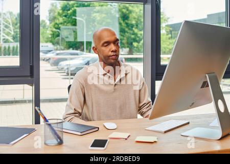 Inklusion, mutiger afroamerikanischer Mann mit Myasthenia gravis, dunkelhäutiger Büroangestellter, der am Schreibtisch sitzt und Computer benutzt, auf den Monitor schaut, Keyboa Stockfoto