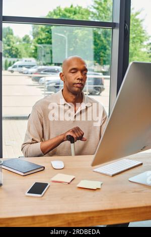 Inklusion, mutiger afroamerikanischer Mann mit Myasthenia gravis, dunkelhäutiger Büroangestellter, der mit einem Gehstock sitzt und Computer benutzt, Monit anschaut Stockfoto