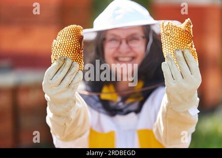 Porträt einer lächelnden Imkerin, die Bienenwachs in einem Schutzanzug im Bienengarten hält Stockfoto