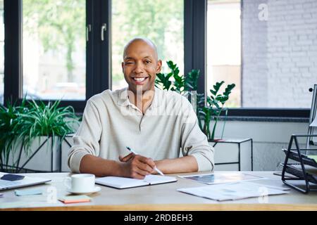 Mann mit Ptosis macht Notizen, glücklicher afroamerikanischer Geschäftsmann, der in die Kamera schaut, glücklicher dunkelhäutiger Büroangestellter mit Myasthenia gravis Krankheit, d Stockfoto